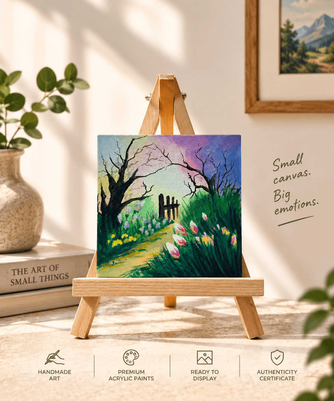 A small square acrylic painting on a wooden easel, depicting a garden path with pink flowers, green grass, and dark tree branches under a purple sky. The scene is styled in a warm, sunlit room with books, a plant, and framed wall art in the background, with text reading 'Small canvas.