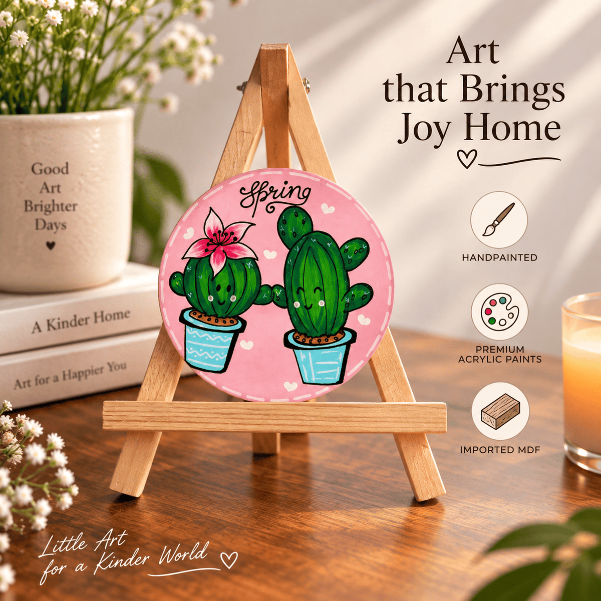 A round hand-painted MDF sign featuring two smiling green cacti on a pink background with the word 'Spring' above them, displayed on a small wooden easel in a styled room scene with books, flowers, and a candle. Text on the image reads 'Art that Brings Joy Home' and 'Little Art for a Kinder World'.