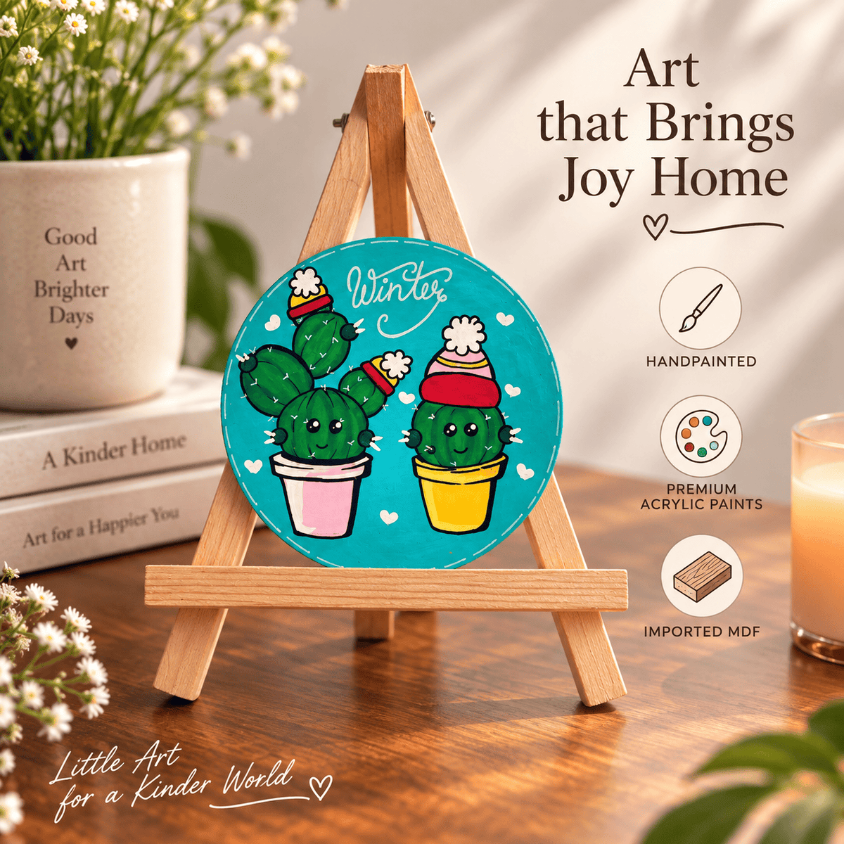 A round handpainted acrylic art panel featuring two cacti wearing winter hats on a turquoise background, displayed on a small wooden easel. The scene is styled on a wooden table with books, a potted plant, and a candle, with promotional text overlays reading 'Art that Brings Joy Home' and 'Little Art for a Kinder World'.