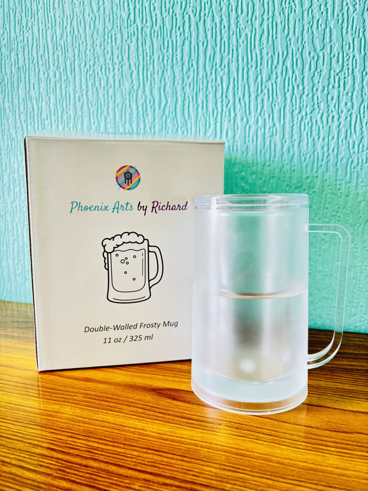 A frosted double-walled mug with a handle sits on a wooden table next to its white product box, which reads 'Phoenix Arts by Richard' and shows a mug illustration. The background is a textured light blue wall.