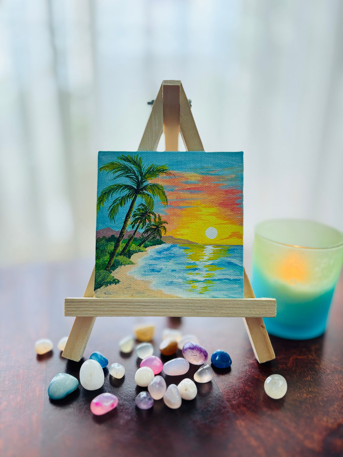 Golden Awakening — a small colorful beach sunset painting with palm trees, blue water, and a yellow-orange sky, displayed on a miniature wooden easel with polished stones and a glowing blue-green candle on a dark wooden table.