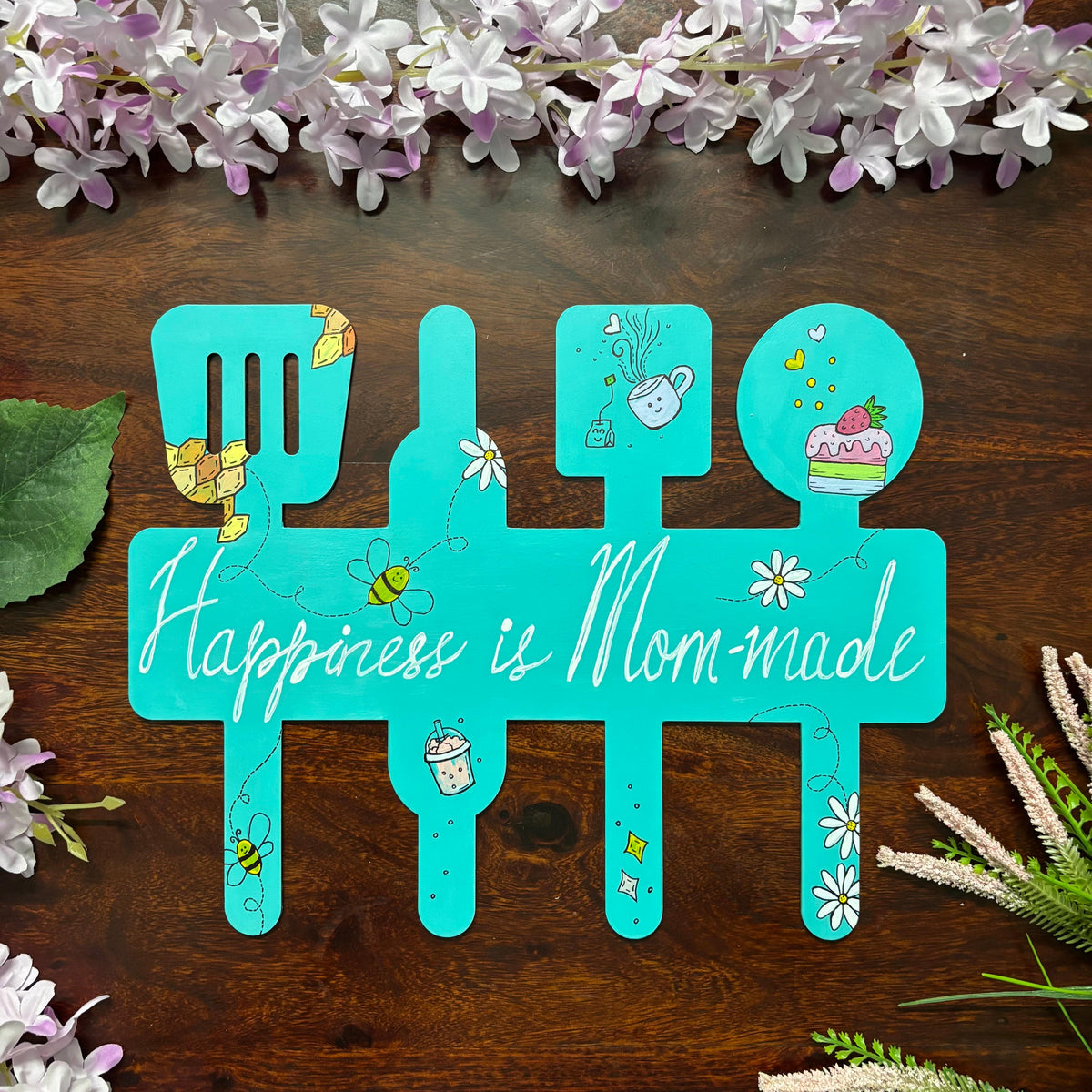 A teal art piece shaped like a kitchen utensil set, featuring the phrase 'Happiness is Mom-made' in white script. The plaque is decorated with small illustrations of flowers, bees, a teapot, a cup, and a cake, and is displayed on a dark wooden surface.