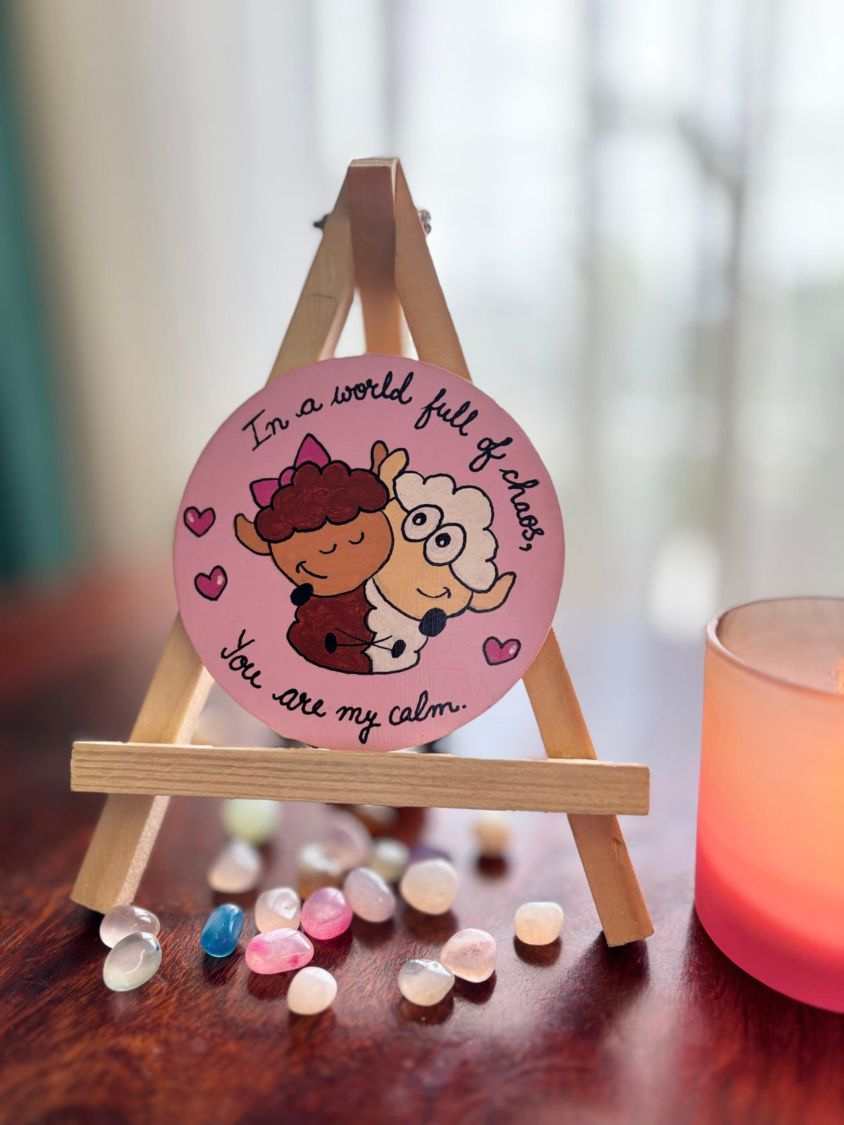A round pink decorative plaque featuring two cartoon sheep hugging with the text 'In a world full of chaos You are my calm', displayed on a small wooden easel on a dark wooden table with scattered glass stones and a glowing pink candle nearby.