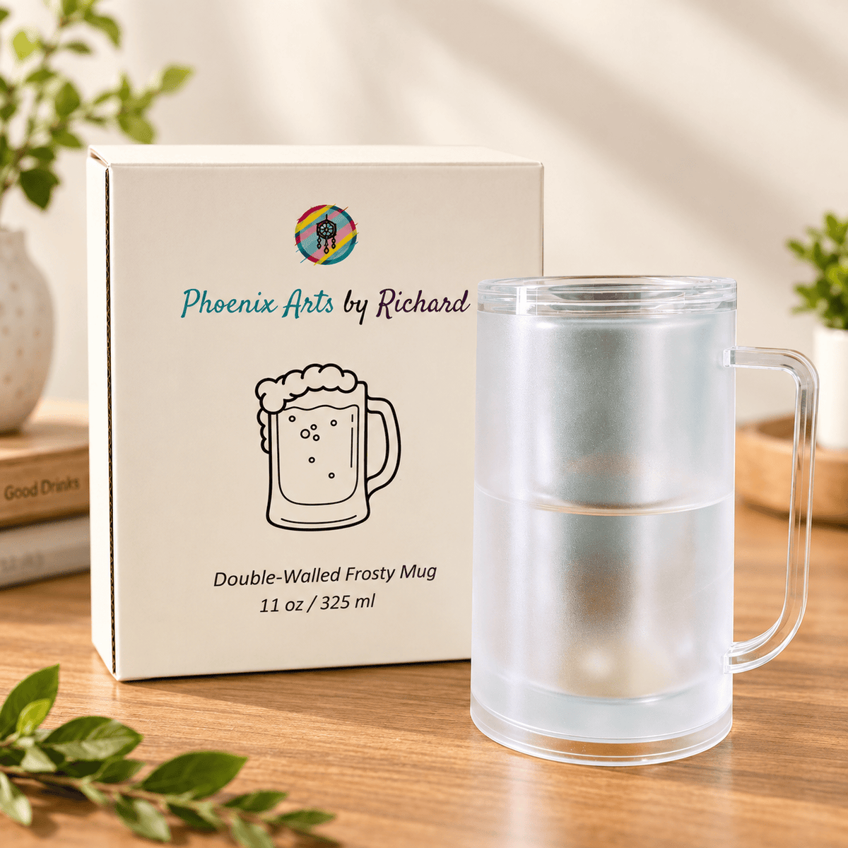 A frosted double-walled frosty mug with a handle sits beside its branded retail box on a wooden surface. The box reads Phoenix Arts by Richard and shows a mug illustration, while the mug itself is transparent frosted glass with a metal infuser visible inside.