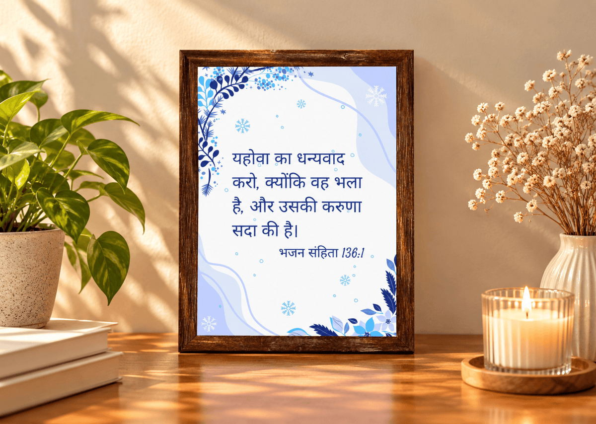 A framed Hindi quote print with a blue and white background and dark blue text, displayed in a dark wood frame on a wooden table. The scene is styled with a potted green plant on the left and dried flowers in a vase with a lit candle on the right, against a warm beige wall with soft shadows.
