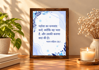 A framed Hindi quote print with a blue and white background and dark blue text, displayed in a dark wood frame on a wooden table. The scene is styled with a potted green plant on the left and dried flowers in a vase with a lit candle on the right, against a warm beige wall with soft shadows.
