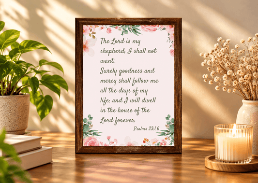 A framed Bible verse print featuring Psalm 23:1-6 with a floral border, displayed on a wooden table in a styled home setting with a potted plant, books, and a lit candle.