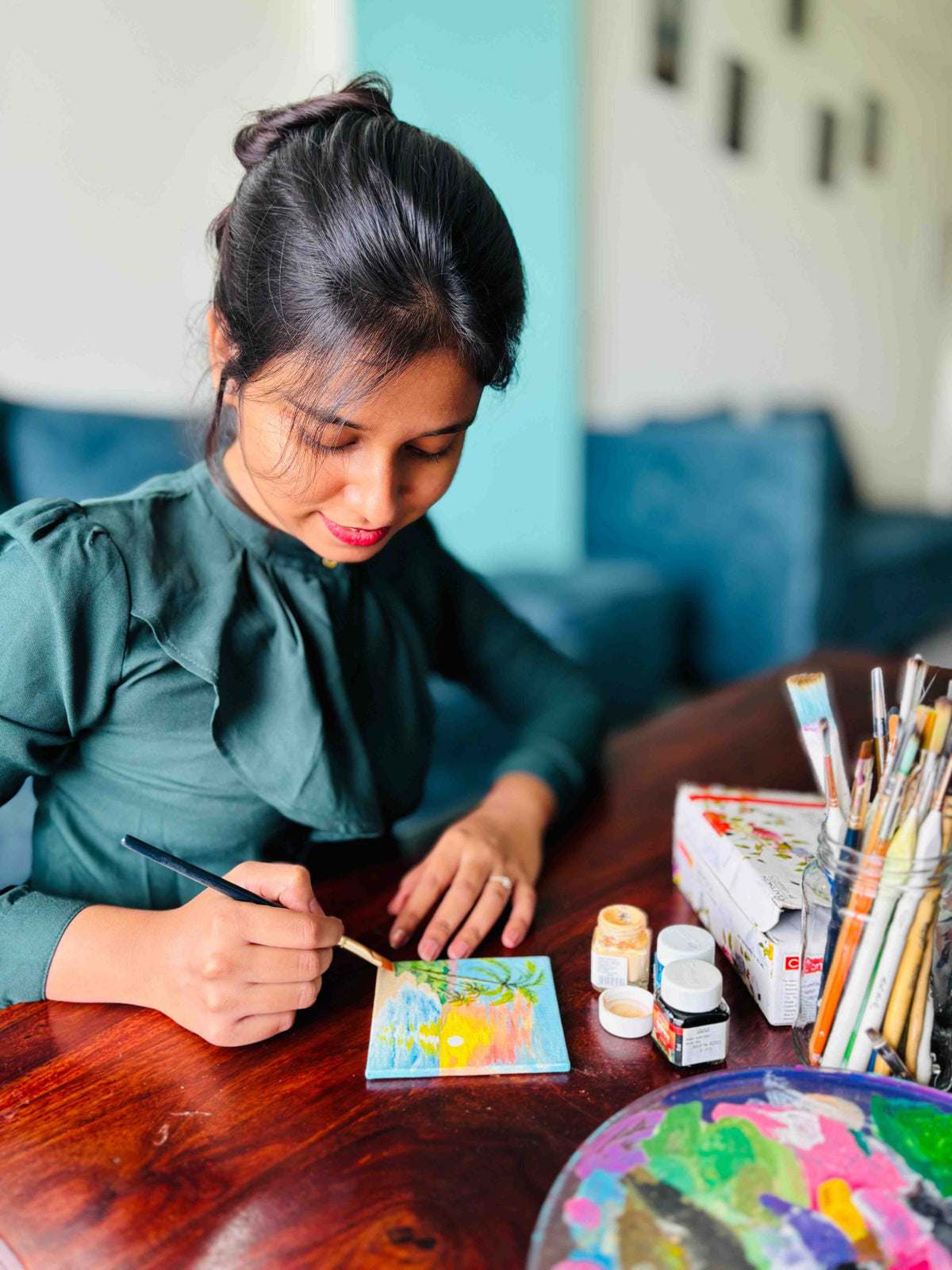 A woman in a dark green blouse painting a small colorful square canvas on a wooden table, with paintbrushes, paint bottles, and a palette nearby in a home studio setting.