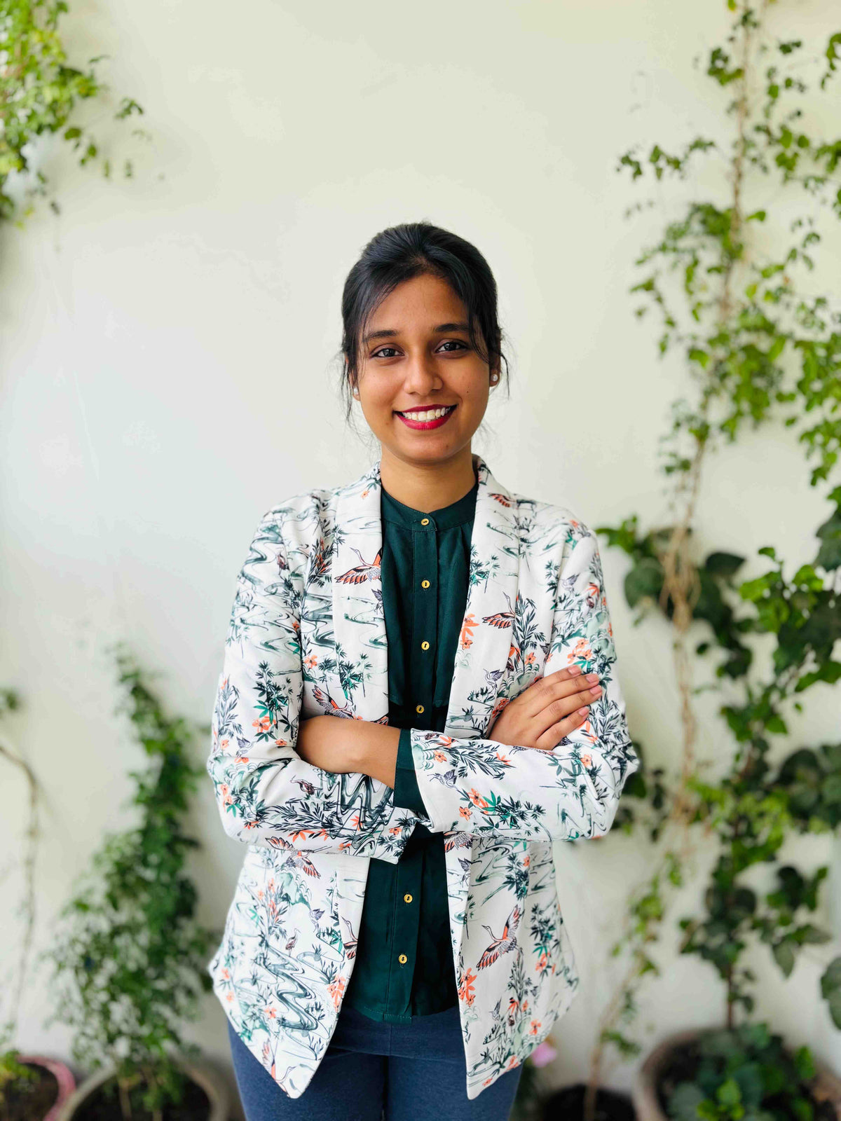 A woman smiling with arms crossed, wearing a white floral blazer over a dark green top and blue jeans, standing in front of a light wall with green plants.
