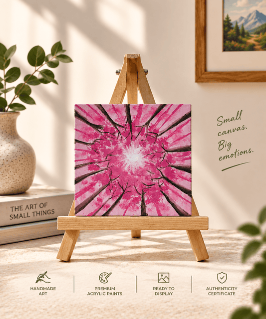 A small square canvas painting displayed on a wooden easel, featuring a bright pink radial floral or tree-bark pattern with a white center. The scene is styled in a warm indoor setting with a vase, books, framed wall art, and text reading 'Small canvas.
