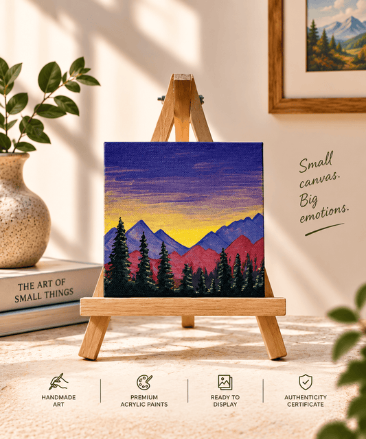 A small square acrylic painting on a wooden easel, depicting colorful mountains and pine trees at sunset. The scene is styled in a warm, cozy room with a vase of green leaves, a framed print, and decorative text on the wall.