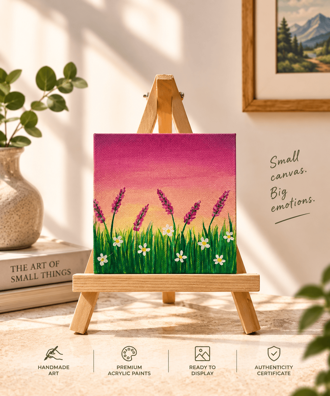 A small canvas painting displayed on a wooden easel in a styled indoor setting. The artwork shows a grassy field with white flowers and purple flowers under a pink and orange sky.