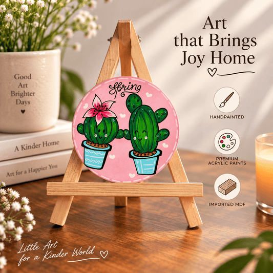 A round hand-painted MDF sign featuring two smiling green cacti on a pink background with the word 'Spring' above them, displayed on a small wooden easel in a styled room scene with books, flowers, and a candle. Text on the image reads 'Art that Brings Joy Home' and 'Little Art for a Kinder World'.