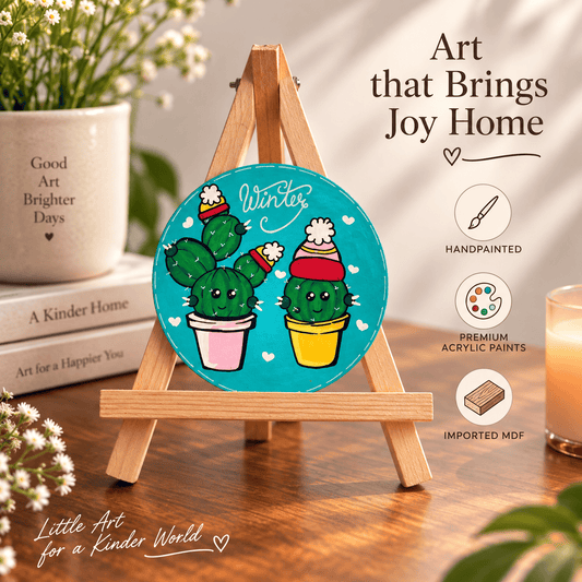 A round handpainted acrylic art panel featuring two cacti wearing winter hats on a turquoise background, displayed on a small wooden easel. The scene is styled on a wooden table with books, a potted plant, and a candle, with promotional text overlays reading 'Art that Brings Joy Home' and 'Little Art for a Kinder World'.