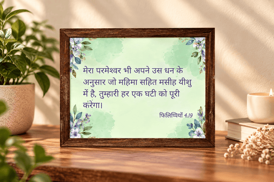 A framed Hindi scripture print with a light green watercolor background and floral corner accents, displayed on a wooden table with a potted plant, candle, and dried flowers in a warm indoor setting.