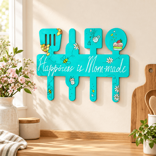 A turquoise kitchen wall sign shaped like utensils with the phrase “Happiness is Mom-made” in white script, decorated with small floral and bee illustrations. The sign is displayed on a wooden countertop in a bright kitchen setting with flowers, a cutting board, and plants.